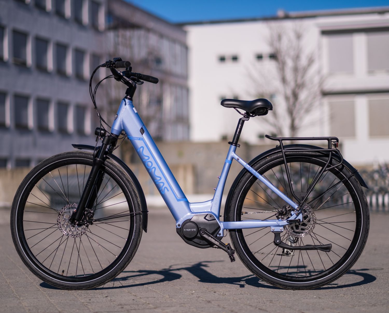 Miljøbilde av en blå elsykkel til dame. Bildet er tatt utendørs på en parkeringsplass med bygninger i bakgrunnen. Det er buet styre og svart sete på sykkelen, med bagasjebrett.